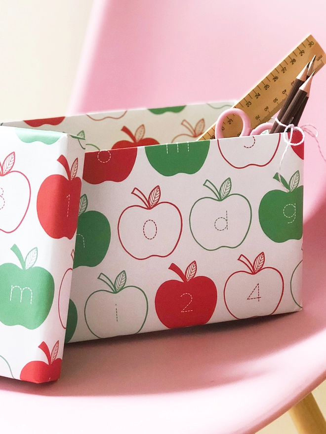 A gift wrapped in red and green apple wrapping paper, with letters and numbers inside each apple, rests on a pink chair.