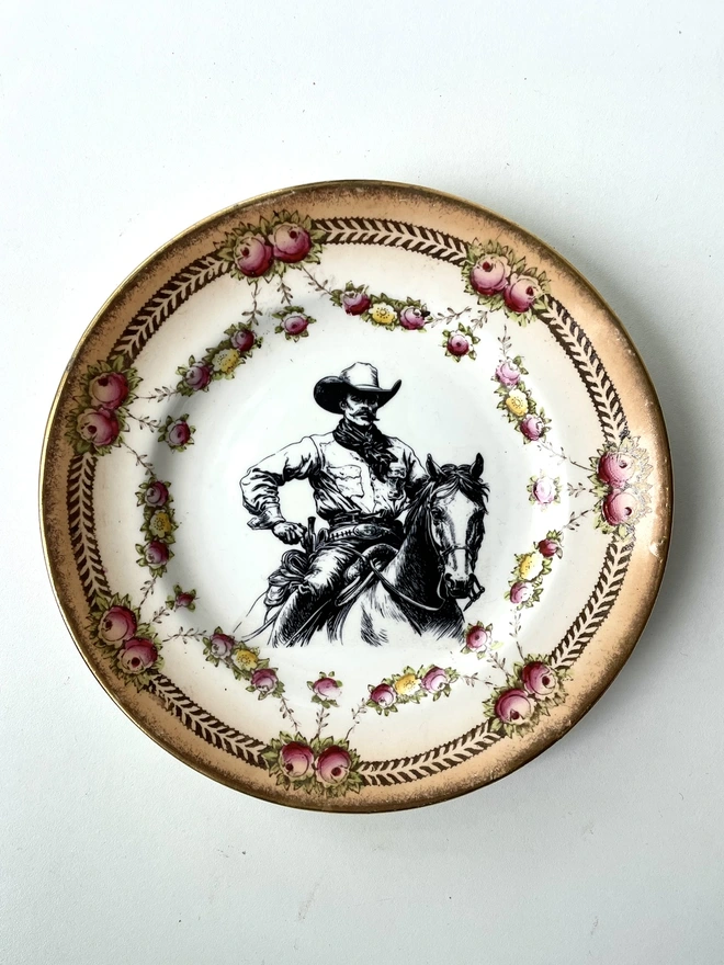 a vintage ceramic china plate with ornate edge in florals and gold, in the centre is a vintage black and white illustration of a handsome cowboy on a horse