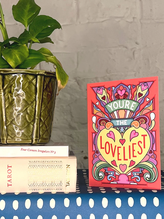 A greetings card with the phrase “You’re the Loveliest” surrounded by an abstract multi-coloured design and lots of hearts, sits on a shelf covered in a blue and white spotted wrapping paper, next to a pot plant sat on top of two books.