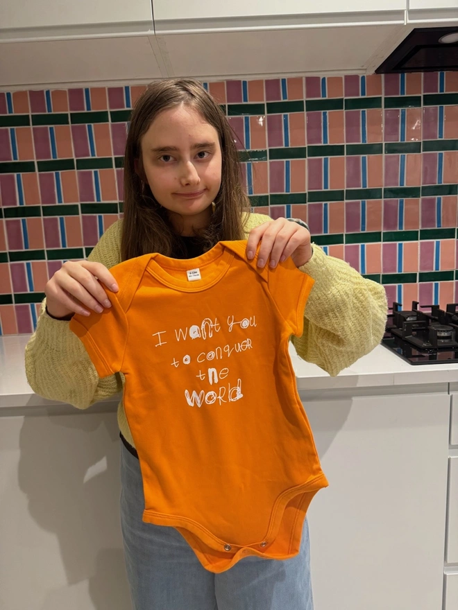 Piper with a cheeky smile holding up an orange baby-sit with her white unique font on it