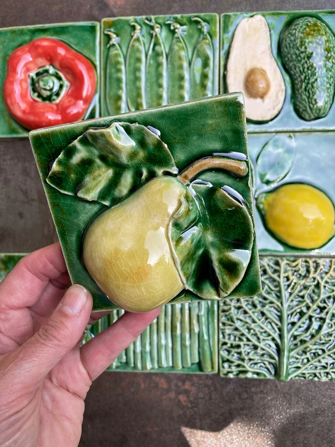 Pear tile - square, 3D, realistic and glossy. The voluptuously shaped pear with leaf and stalk has a lush green glaze. Other fruit and vegetable tiles in the series are on display in the background: lemon, fig, savoy cabbage, red capsicum pepper, mange tout, avocado, garlic, asparagus.