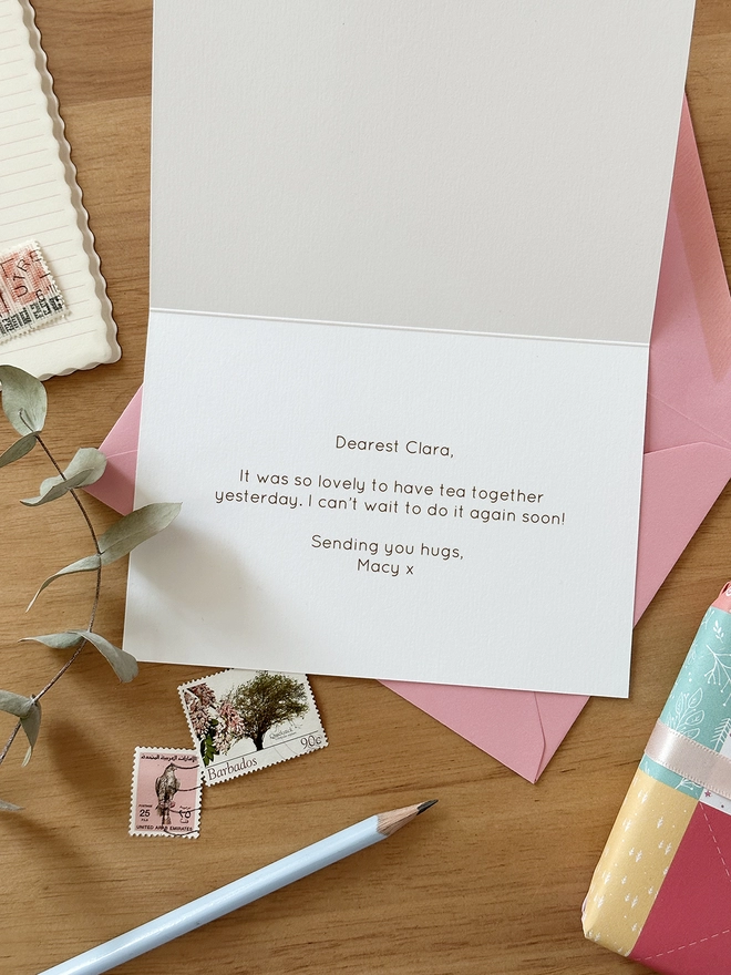 A greetings card with a printed message inside lays on a pink envelope on a wooden desk.