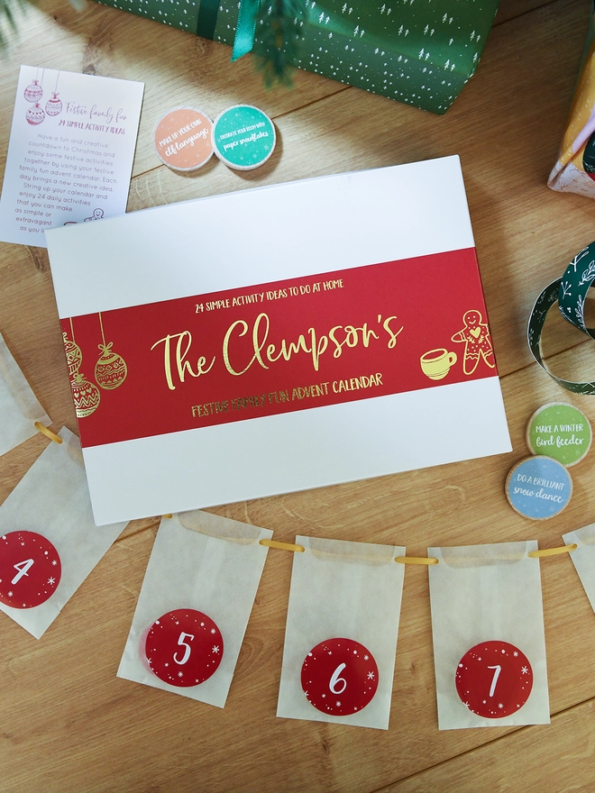 A white advent calendar box with a red sleeve lays on a wooden floor with white numbered envelopes beside it.