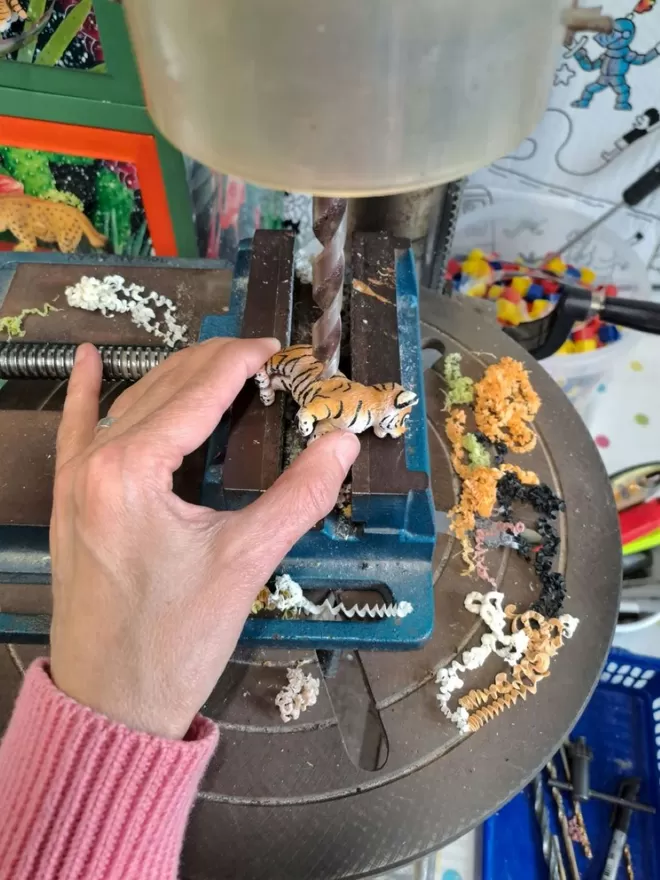 A hand holds a small plastic tiger on a clamp on the table of a pillar drill. A chest of safari style drawers can be seen in the background with animal knobs attached. The tiger is being drilled by Candy Queen Designs and made into a decorative dimmer switch for a child's nursery. 