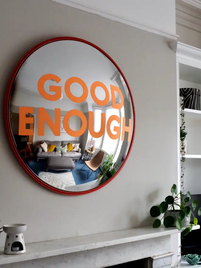 Round convex mirror with 'Good Enough' hand painted in orange in a red round frame, above a cream fireplace surrounded by house plants