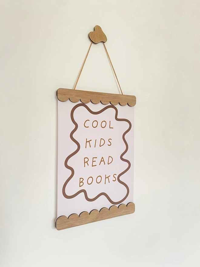 A wooden magnetic picture frame, strung with vegan suede. Hung on a wavy wooden wall dot. Displaying a colourful stripy kids print that says cool kids read books 