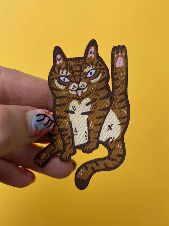 Illustrated cat licking themselves in the form of a woven iron-on patch 