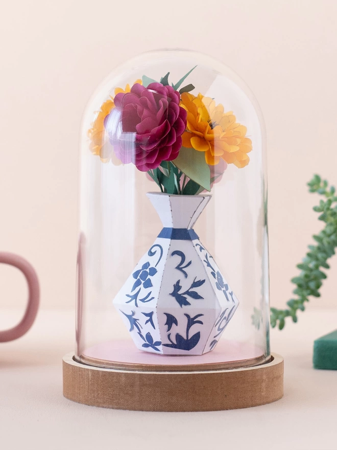 a bunch of paper flowers in a white and blue vase on a plastic dome . This is a craft kit by My papercut Forest