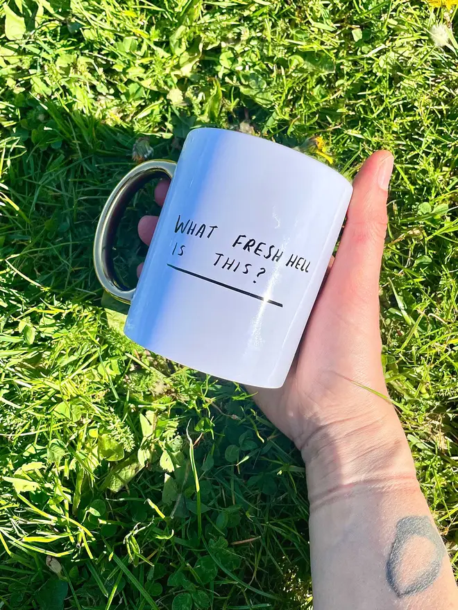 Holding mug against grass background. Mug reads 'what fresh hell is this?'