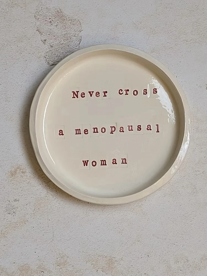 'Never Cross A Menopausal Woman' Ceramic Dish', a ceramic dish sitting on a grey surface. It is inscribed with a message in red writing. 