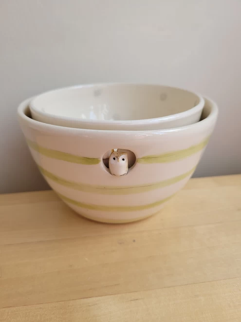 Two pottery bowls on a wooden counter top the smaller bowl is stacked inside the larger bowl which is green and off white stripes with a ceramic owl figure attached inside a cut out