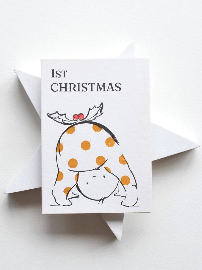 Christmas greeting card resting on a white star backdrop.  Card features a baby in spotty outfit peering through legs with a holly sprig on top to resemble a christmas pudding. Wording at the top says '1st Christmas'. 