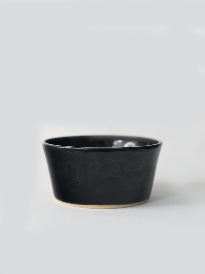 A single hand-thrown ceramic breakfast bowl in jet black, with a glossy glaze showing subtle variation in tone, a gently flared form and unglazed natural clay base, photographed against a plain light grey background.