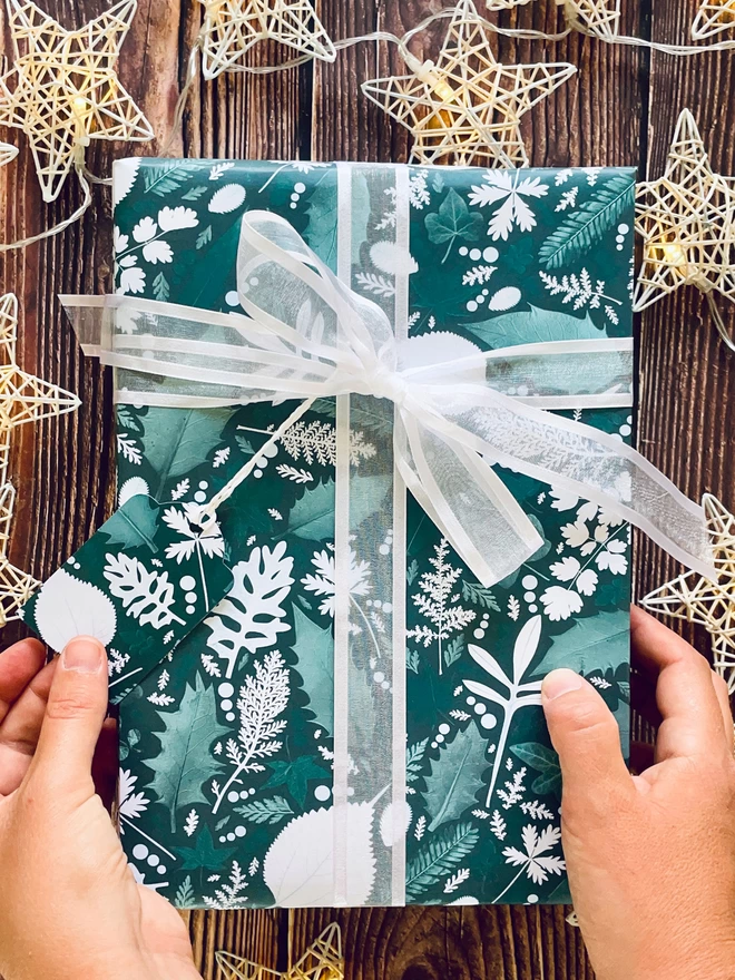 Hands holding Christmas gift wrapped in green pressed leaf wrapping paper with Holly and Ivy design