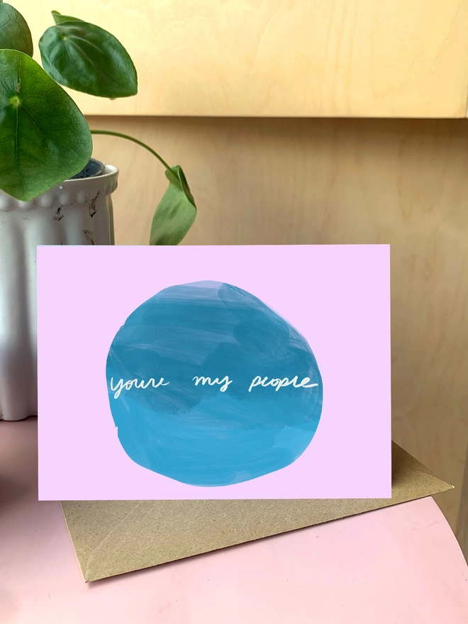 Purple greeting card with blue circle, within it reads 'you're my people'. Card sits on table with plant.