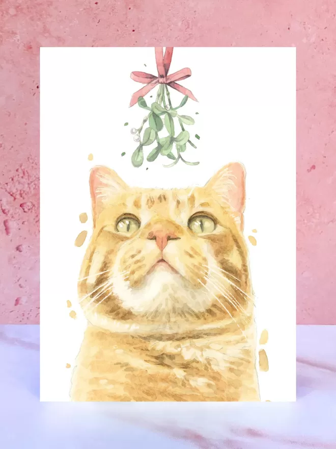 A Christmas card featuring a hand painted design of a Ginger and White Cat, stood upright on a marble surface.