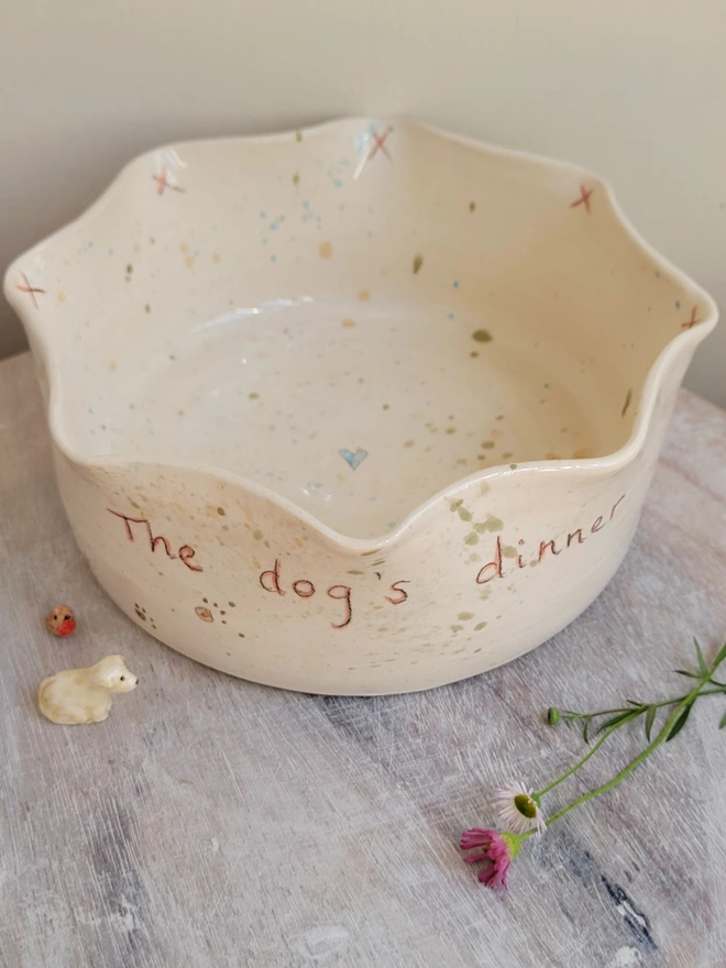 ivory ceramic dog bowl with 'the dog's dinner' written on the side with brown green and blue splatters and pawprints with a blue heart in the centre of the bowl