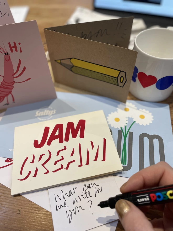 An array of colourful bold greetings card are laid out with a Devon mug behind. A loose energetic handwritten message writes What Can We Write for You? Hand is poised with a pen. Cards around read, Jam Cream typography, the word Mum with flowers poking out, A prawn says hi!, a graphic yellow pencil. All exuding joy and life!