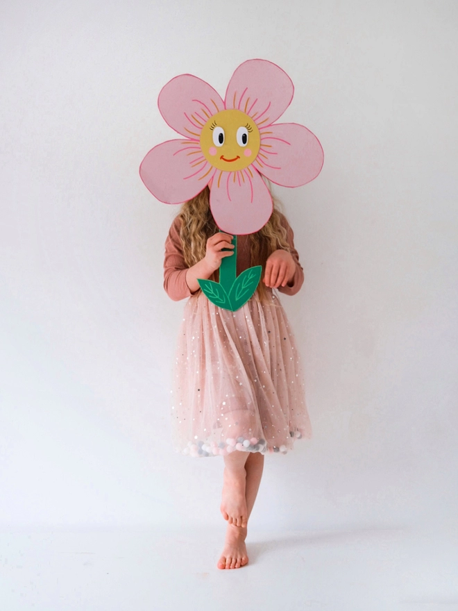 A young girl posing holding a giant cut out flower covering her face while wearing a pink pom pom tutu dress
