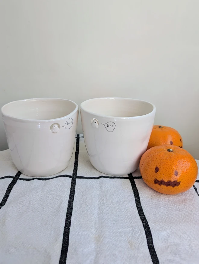 two small ghost cups with finger dents in white handmade ceramic with a tiny sculpted ghost in a cut out near the top of the beakers with a hand painted bubble with the word boo