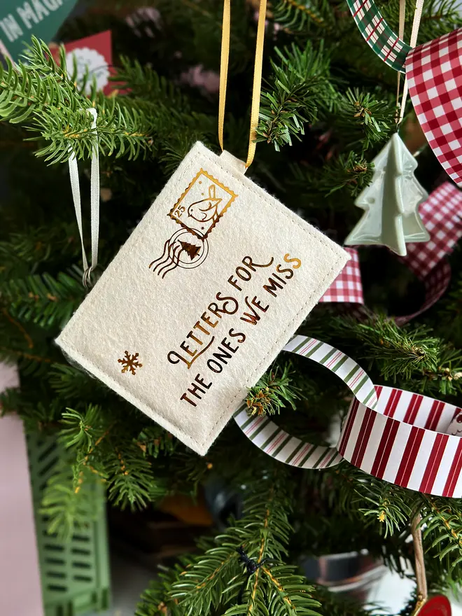 A handmade ivory felt envelope decoration with the words 'Letters for the ones we miss' in gold hangs in a Christmas tree.