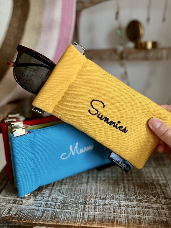 Hand holding a bright yellow cotton sunglasses pouch with 'Sunnies' embroidered on it. Other colours of glasses holders in the background.