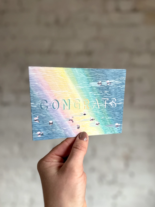 a greetings card featuring an illustration of of flamingos in a lake with a rainbow reflection with the text “CONGRATS”