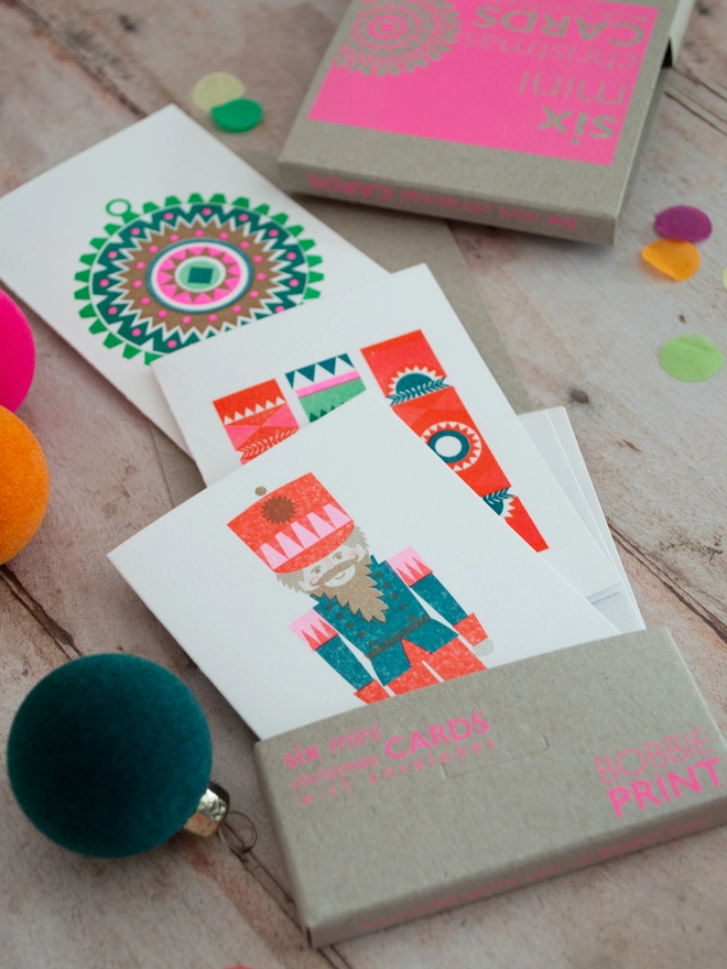 A boxed set of mini Christmas cards lies open on a wooden table, three designs include a retro geometric bauble design, crackers and a festive nutcracker. Small brightly coloured baubles are scattered around the box of cards.