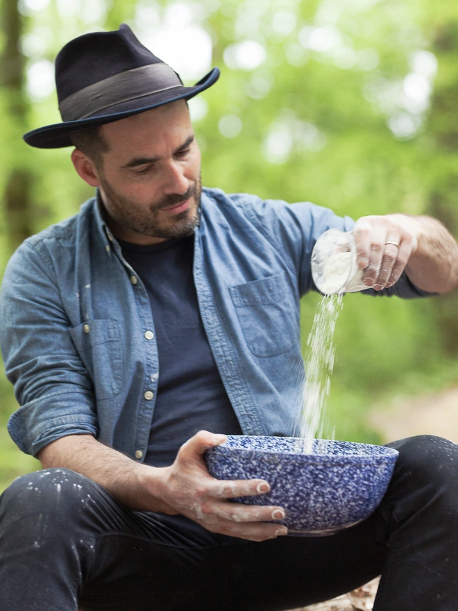 Man sitting on tree trunk with bowl in one hand and shaking a cup of flour into it with his other hand