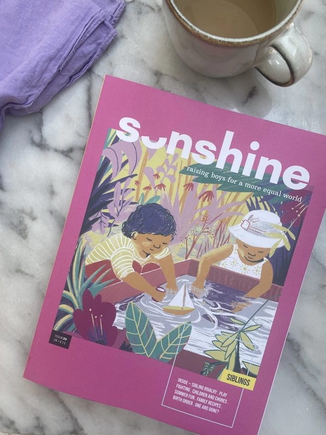 Sonshine Magazine Issue 20 , a pink magazine with an illustrted image of two children playing in water together.. The magazein is lying on a marble table top with a cup of tee and a purple napkin next to it. 