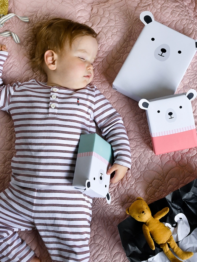 A baby wearing red and white striped pyjamas is sleeping on a pink quilt with several gifts wrapped as polar bears laying beside.