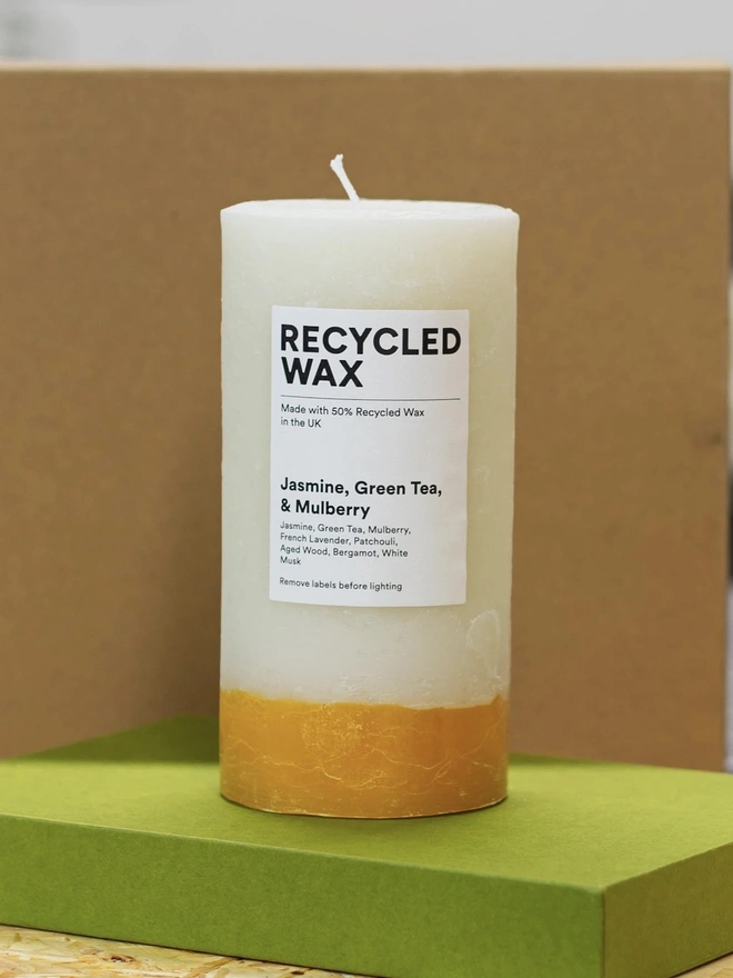 Jasmine, Green Tea & Mulberry Candle (Set Of 1 Or 3 Wicks). a taller jasmine, green tea and mulberry candle stands on a green box against a brown background, showing its single wick and cream wax with a golden yellow base. a white label reads "recycled wax, made with 50% recycled wax in the uk", followed by the scent name and note list of jasmine, green tea, mulberry, french lavender, patchouli, aged wood, bergamot and white musk. the candle's two-tone colouring and visible drip texture give it a rustic, hand-poured look.