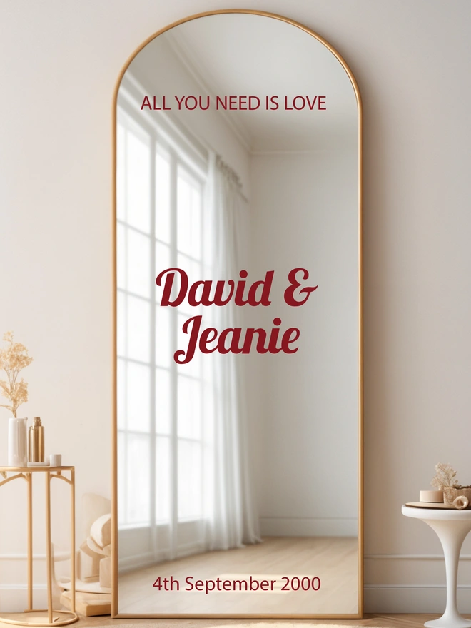 Large mirror leaning against a white wall with a table next to it, on the mirror is the wording All you need is love, David and Jeanie and the date of the wedding in Burgundy lettering.