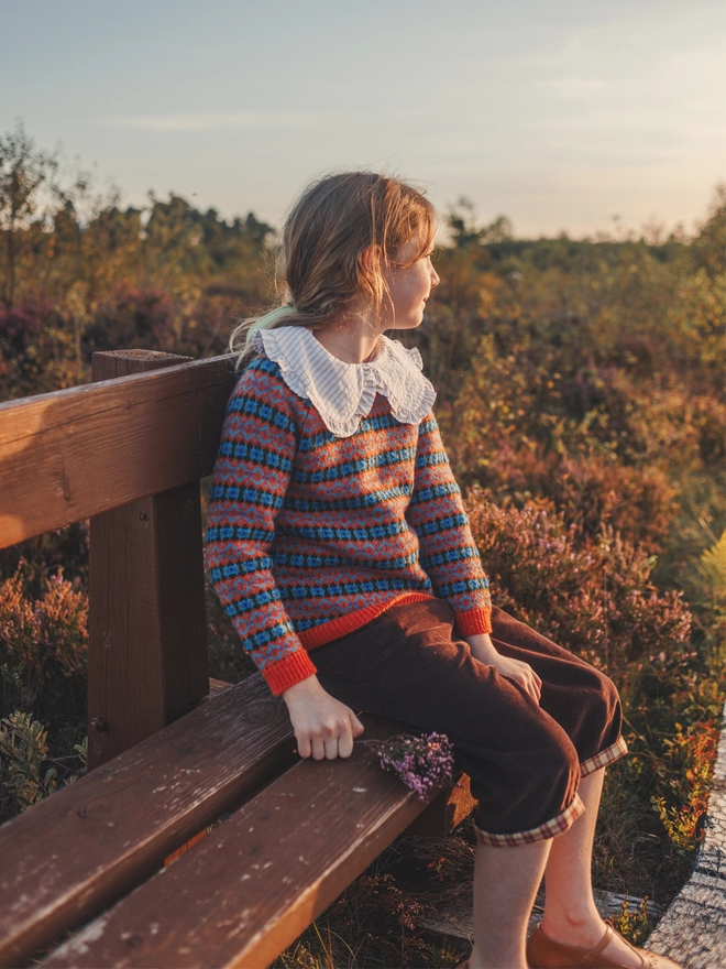 A girl in an orange fairisle jumper and brown cropped trousers sits on a bench