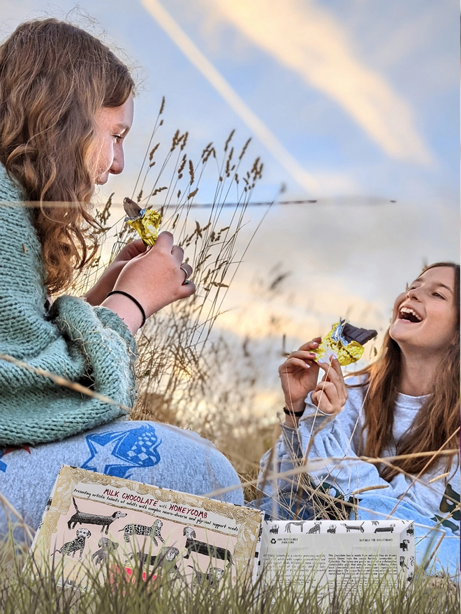 Two young happy girls in a field enjoying eating charity milk chocolate bars with dog on the wrapper
