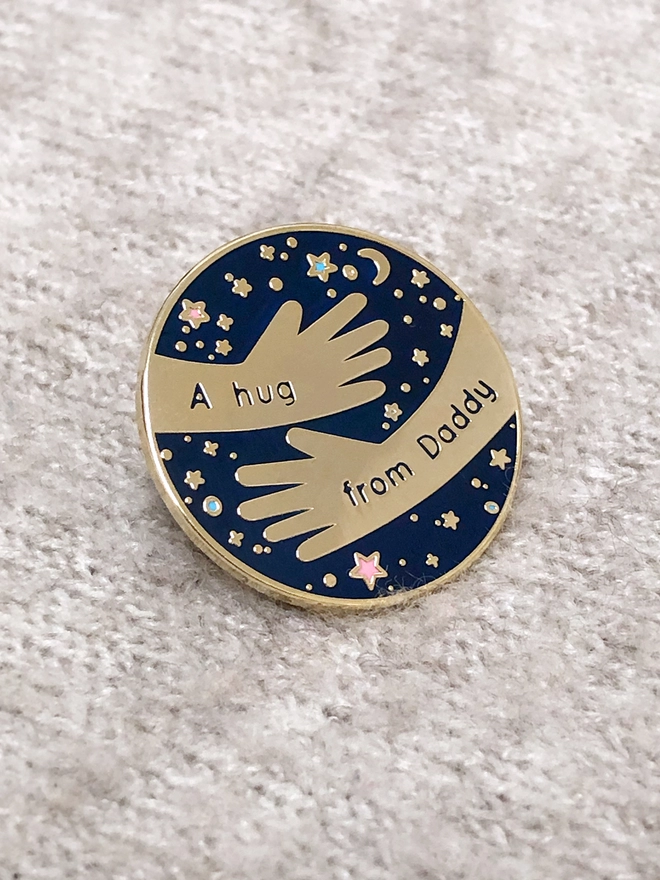 A navy blue and gold enamel pin badge with a hugging arms design and the words "A hug from Daddy" is pinned to a beige blanket.