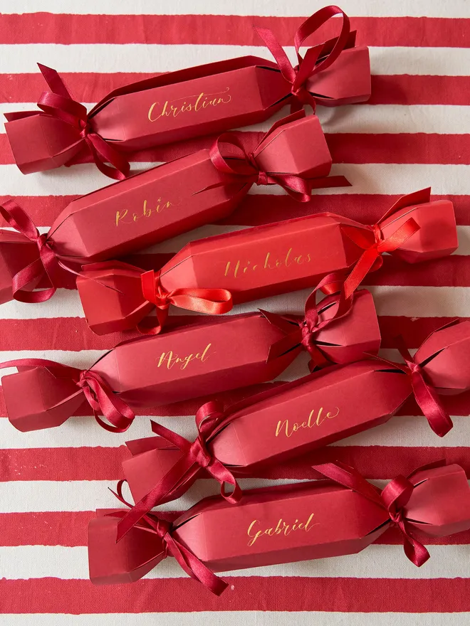 A set of 6 personalised Christmas crackers in red with gold calligraphy with matching ribbon, atop a red and white striped tablecloth