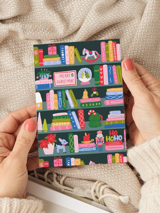 A picture of someone holding a Christmas card in their hands with a Christmas bookshelf illustration.