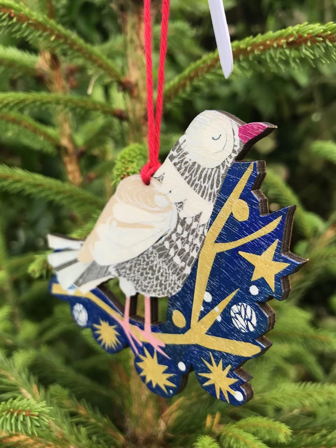 Illustrated peace dove standing on a gold star-spangled twig on a blue backround. The cut-out decoration hnags against the fresh green of a fir tree Christmas tree.