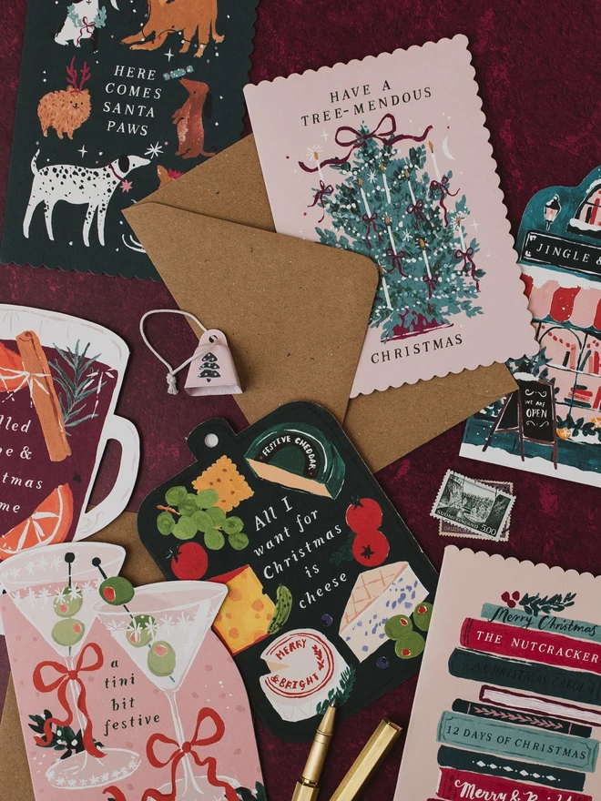 Sister Paper Co Christmas cards laid out on a deep red background, including Santa Paws, Tree-mendous, Cheeseboard, Mulled Wine and Tini Bit Festive