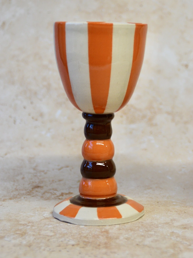 Handmade ceramic wine cup with vertical orange and cream stripes and a stacked stem in brown and orange glaze, photographed on a neutral background.