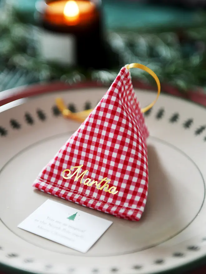 A red gingham fabric fortune cookie with gold personalised name “Martha” sits on a Christmas dinner plate, ready as a handmade festive place setting with a hidden Christmas wish card beneath.