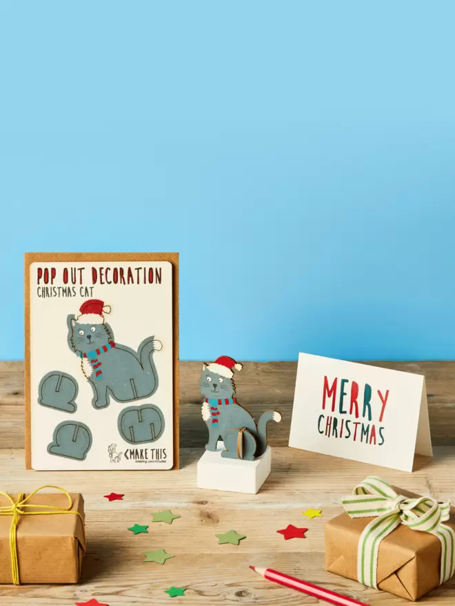 Pop out laser-cut black cat Christmas decoration and Merry Christmas card and brown kraft envelope on top of a wooden desk in front of a sky blue coloured background