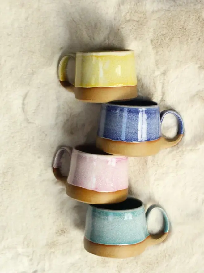 Four handmade ceramic cups with pastel glazes—blue, yellow, teal, and pink—arranged in a line on a soft cream-colored fur surface.