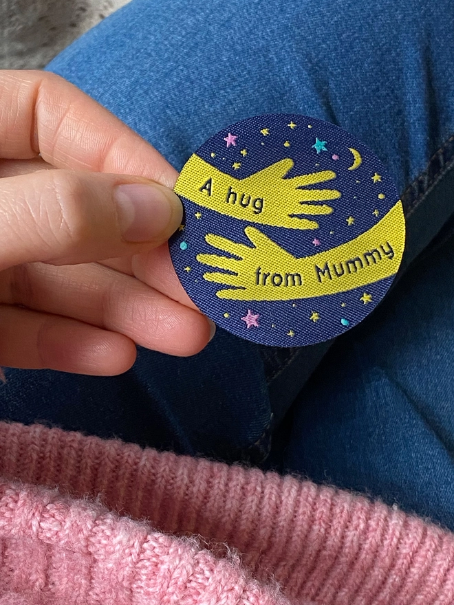A navy blue and yellow woven patch is being held. The design is two arms in a hug shape with the words "A hug from Mummy" along them. 