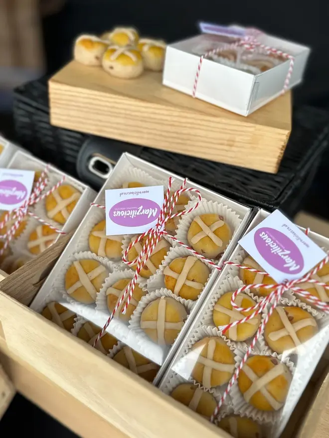 Marzipan Hot Cross Buns (Box Of 6 Or 9), groups of marzipan hot cross buns packaged in white boxes. In the background are more marzipan hot cross buns. 
