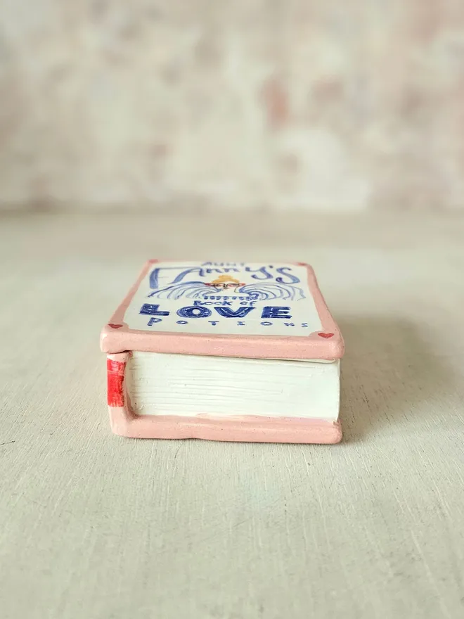  A charming ceramic trinket box designed to look like a vintage book, painted in shades of pink and red. The cover features the title "Aunt Fanny’s Book of Love Potions" in a playful, illustrated style. With its book-like shape and whimsical design, this decorative piece adds a touch of romance and mystery while offering a secret spot to store small treasures.