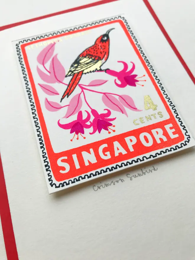 Bird Stamp Card Singapore/ Crimson Sunbird, a white card with a colourful bird design in the centre above a red envelope on top of a white surface. 
