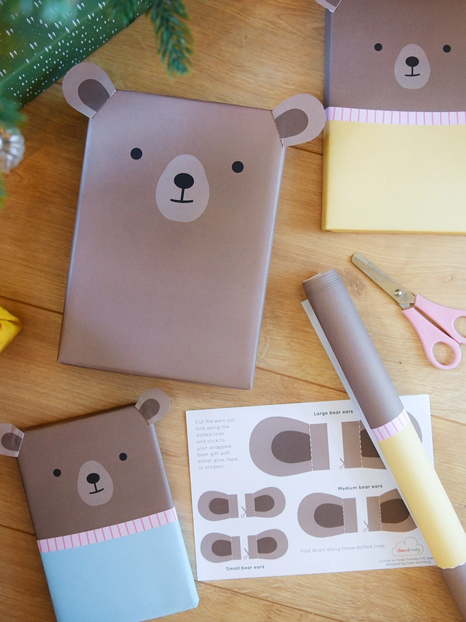 Three gifts wrapped as brown bears lay on a wooden floor beside a sheet of wrapping paper and scissors.