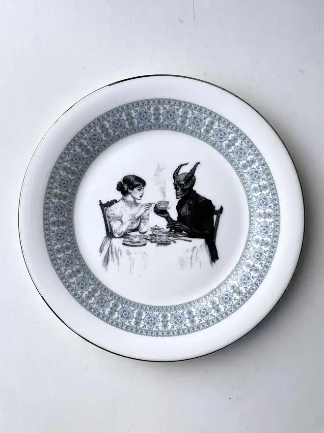 a vintage plate with an ornate edge, in the centre is a vintage illustration of a victorian lady drinking tea with the devil